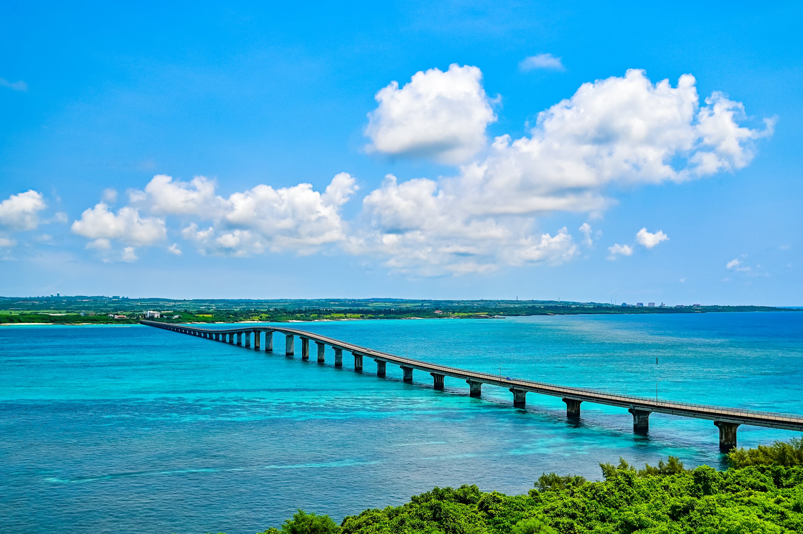 A long, low bridge crossing turquoise shallows to a flat green island under white cloud