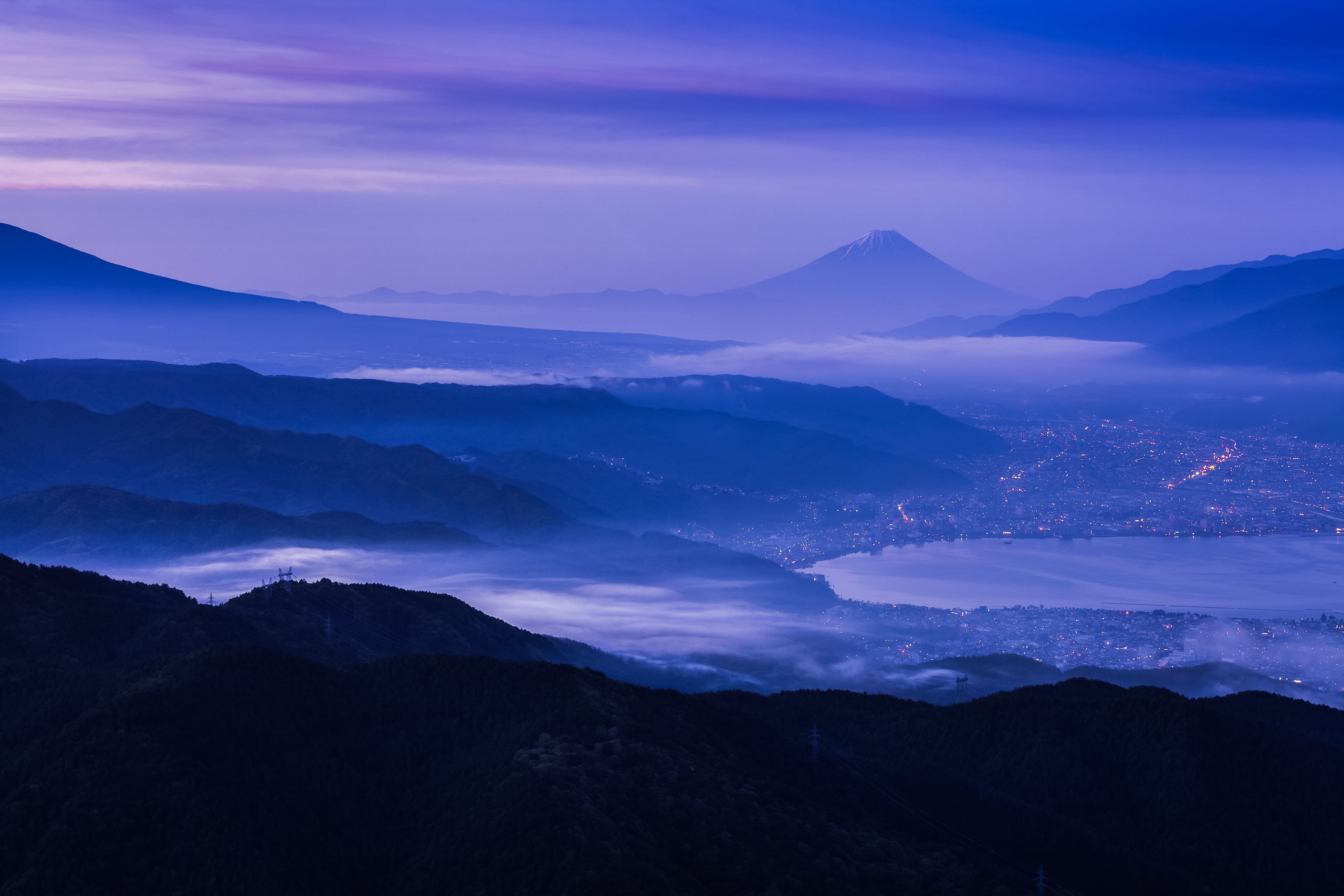 Layered ridges of mountain at blue hour beyond a lakeside town, the cone of Mount Fuji small on the far horizon