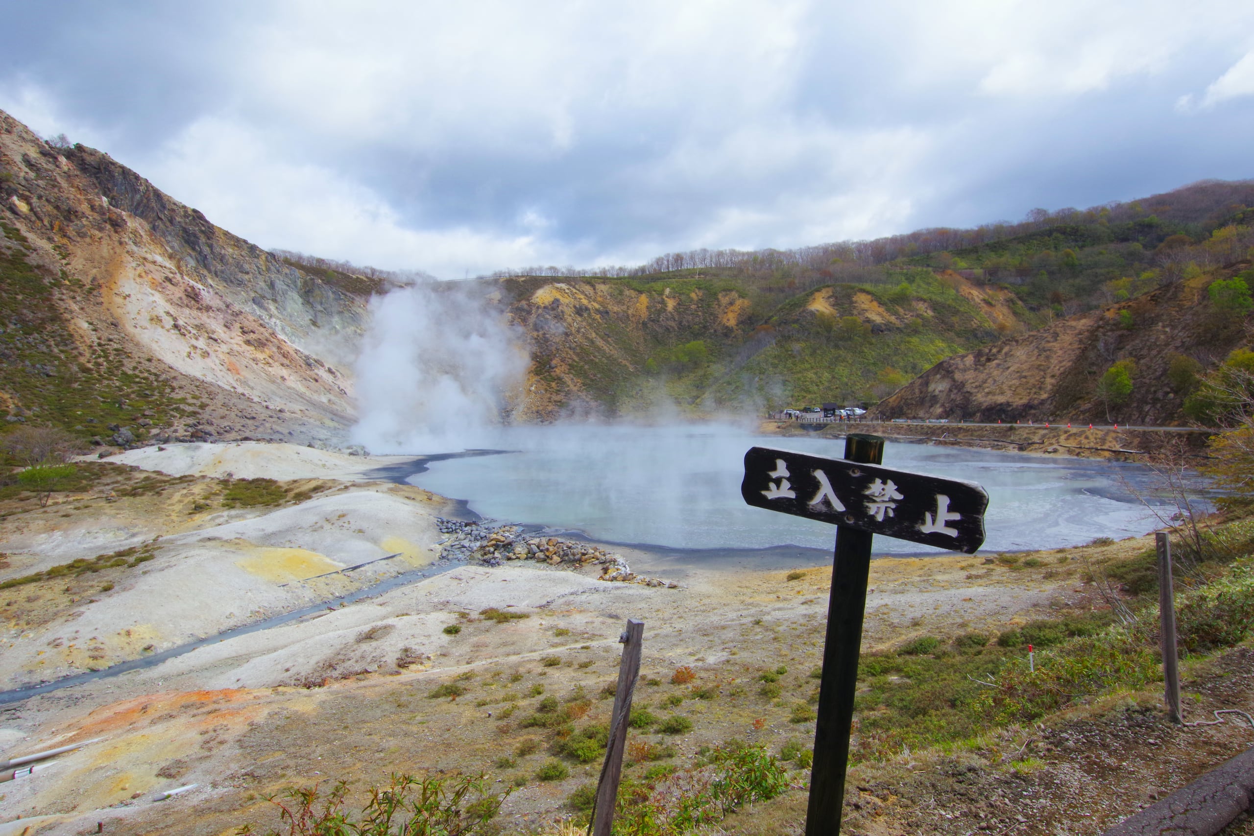 Steam rising from the grey-green water of the Oyunuma crater pond beneath a forested slope