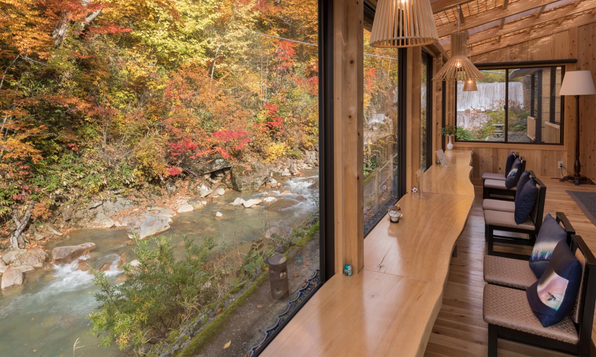 A long timber lounge with low chairs at a window onto a river and autumn trees, Taenoyu