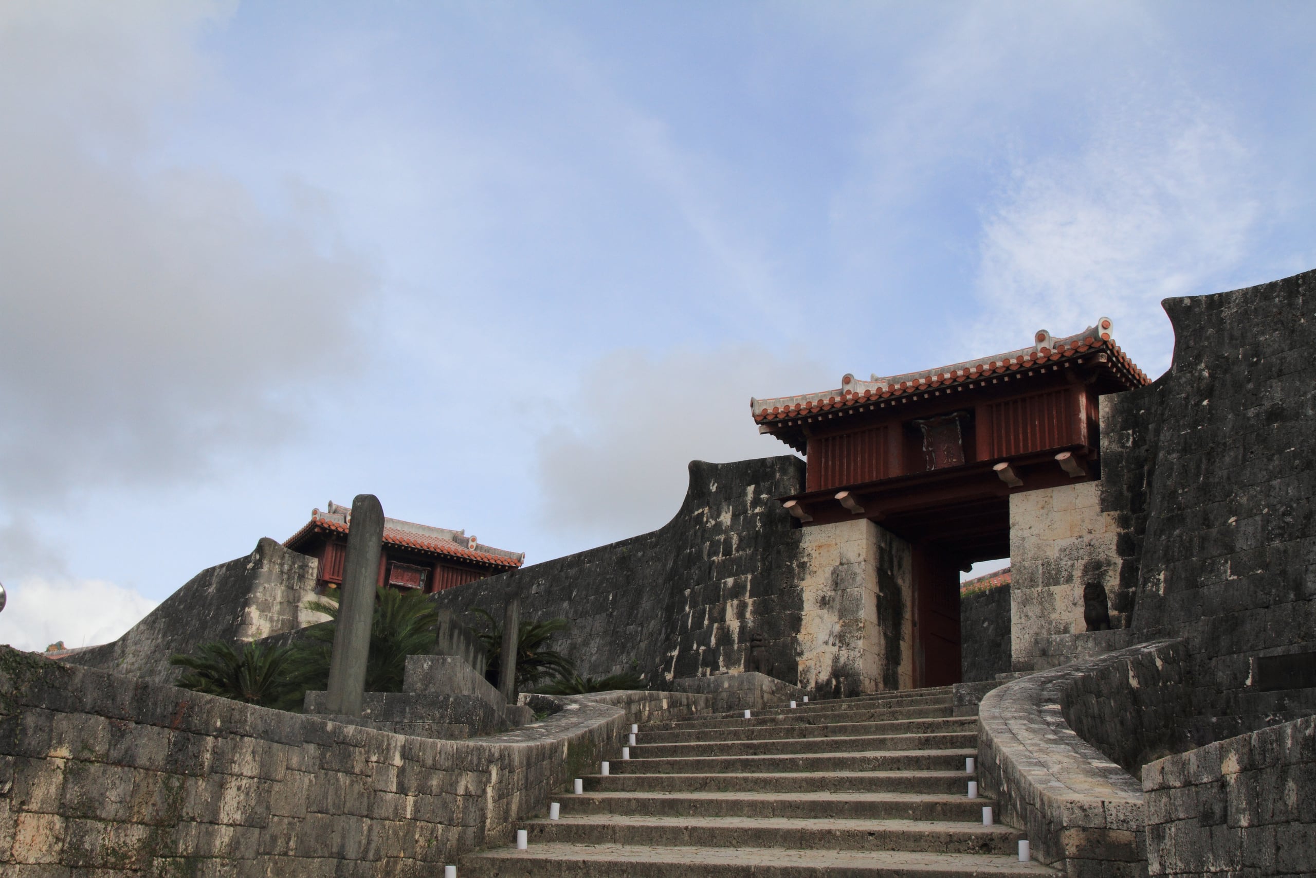 A red-lacquered gate with a white-tiled roof set into curved stone walls at the top of a stair