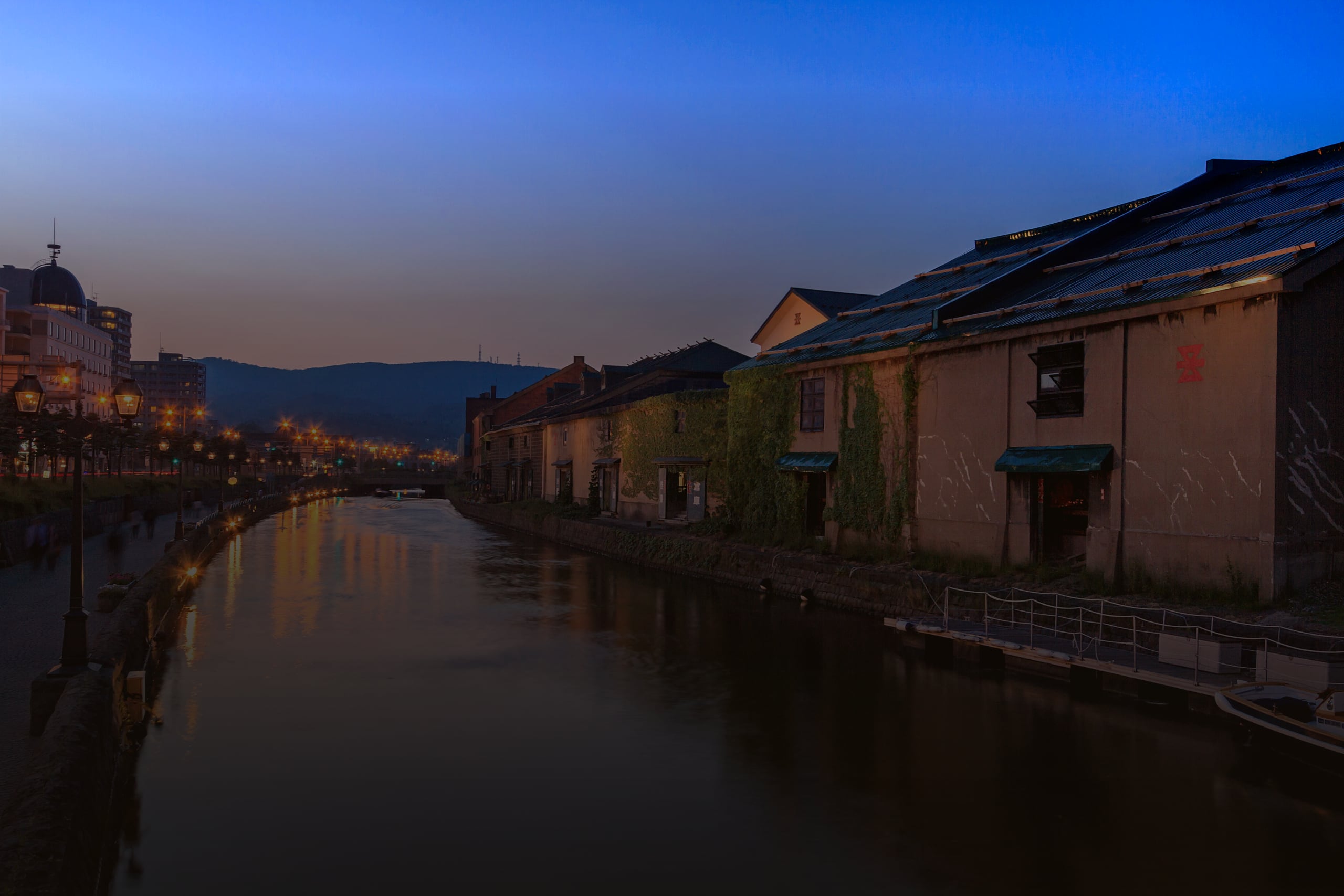 The Otaru canal at blue hour, gas lamps lit along the towpath and the stone warehouses reflected in the water