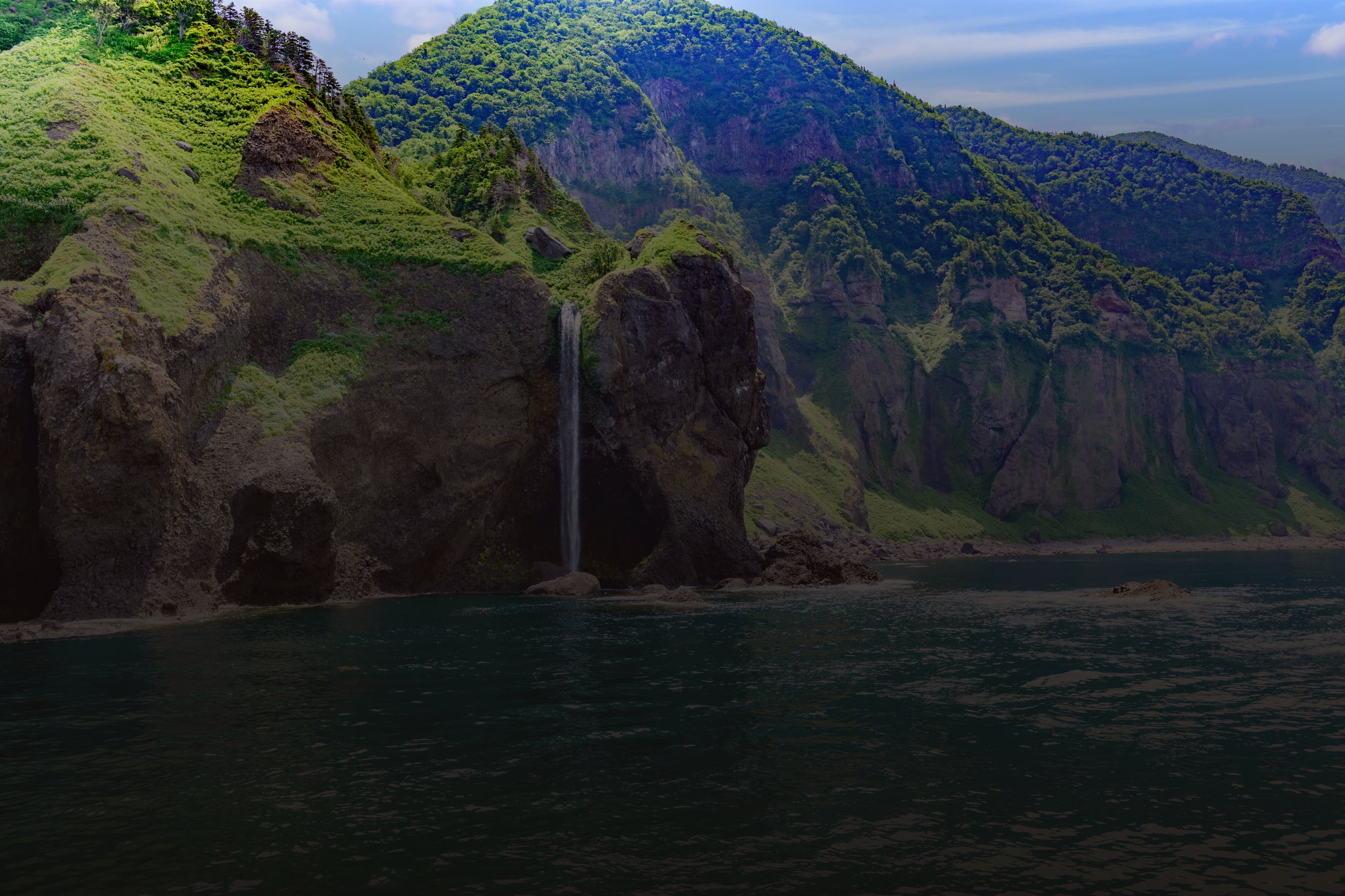 A waterfall dropping off a green basalt cliff straight into the blue sea on the Shiretoko coast