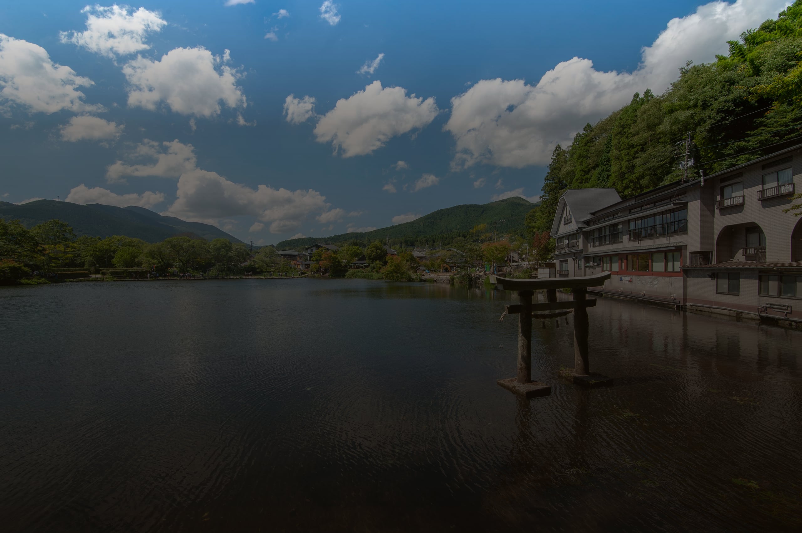 A stone torii standing in the still water of Lake Kinrin, inns on the shore and green hills beyond