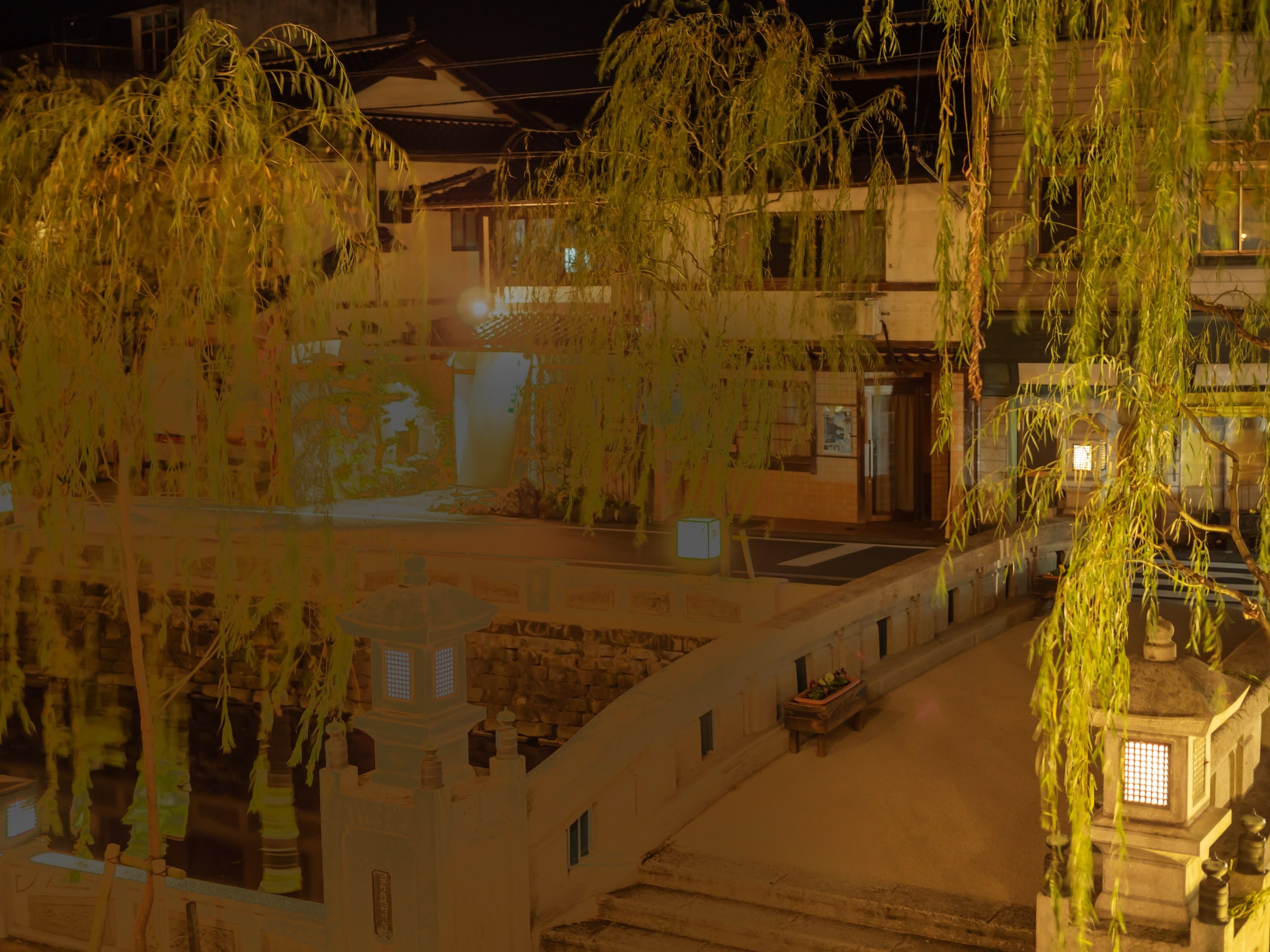 A stone bridge with lantern posts over the willow-lined canal of Kinosaki Onsen at night, the lit windows of the town behind
