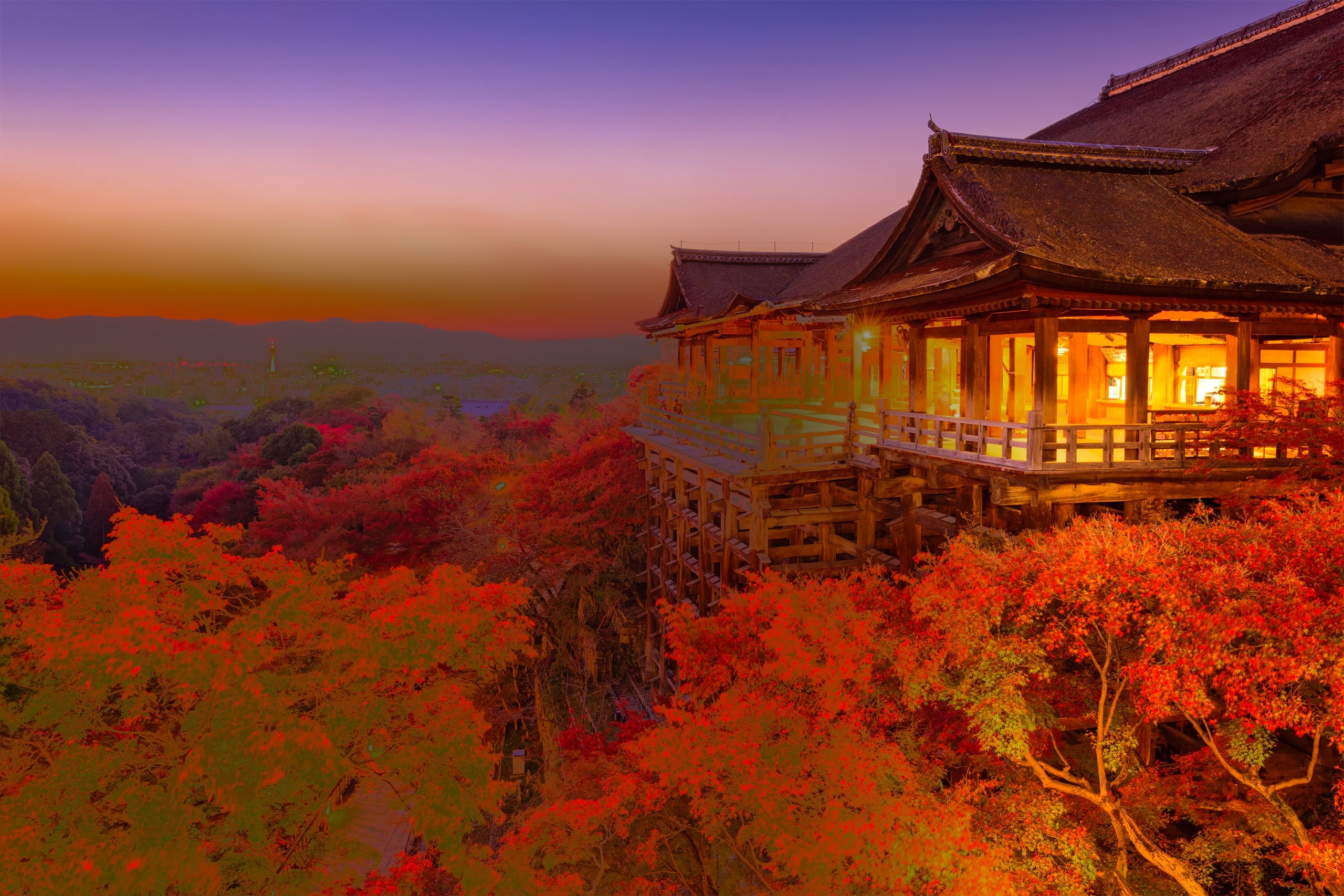 The wooden stage of Kiyomizu-dera lit at dusk on its tall pillars, red maples below and the lights of Kyoto beyond