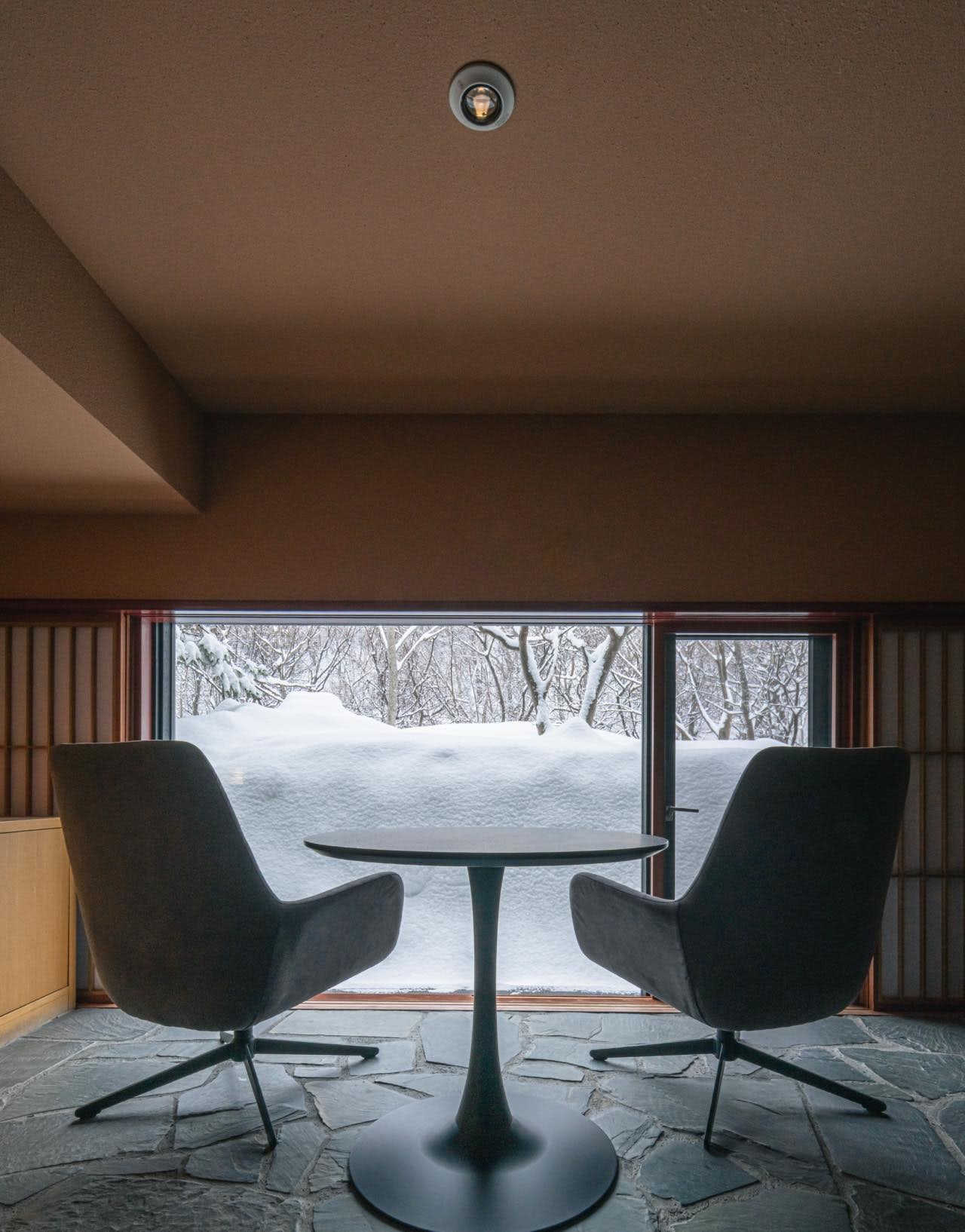 Two chairs and a round table before a wide window onto snow-covered trees, in a room at Kuramure