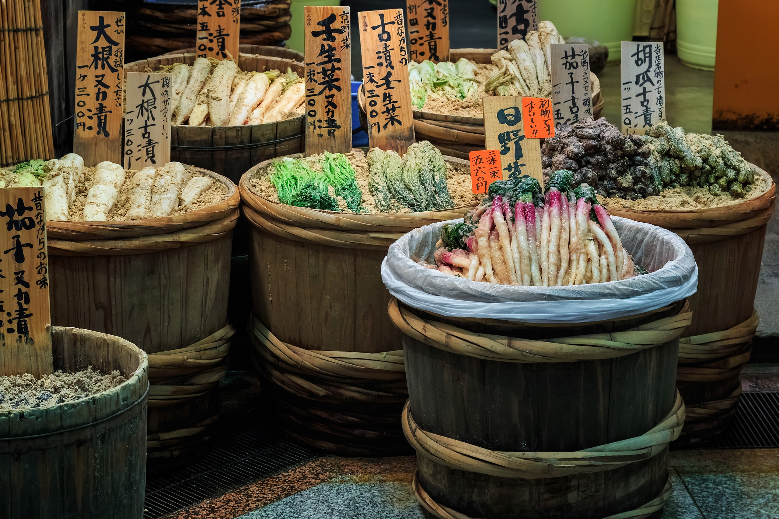Pickled vegetables piled in wooden tubs at a market stall, each tub labelled by hand