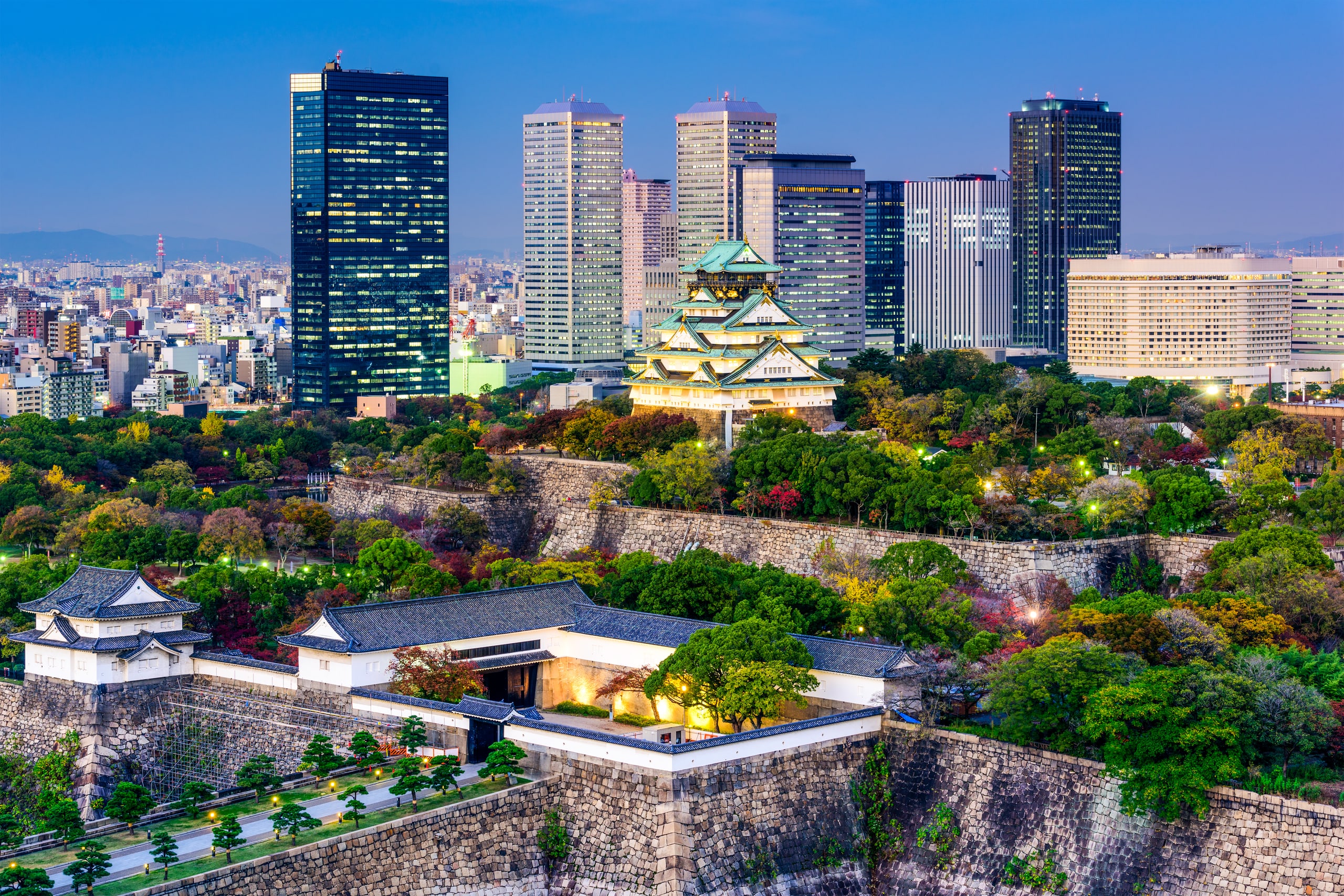The keep of Osaka Castle above its stone walls and turret, with the towers of the business district behind, at dusk
