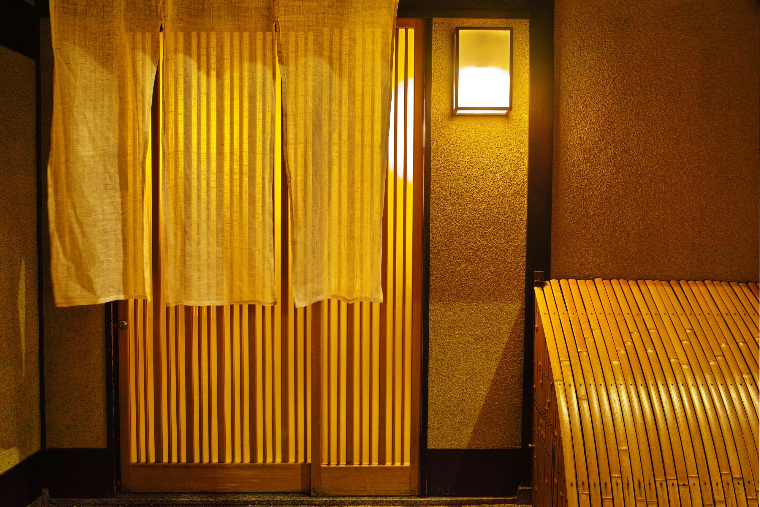 The entrance of a restaurant at night, a cream noren curtain hanging over the lit timber of the doorway