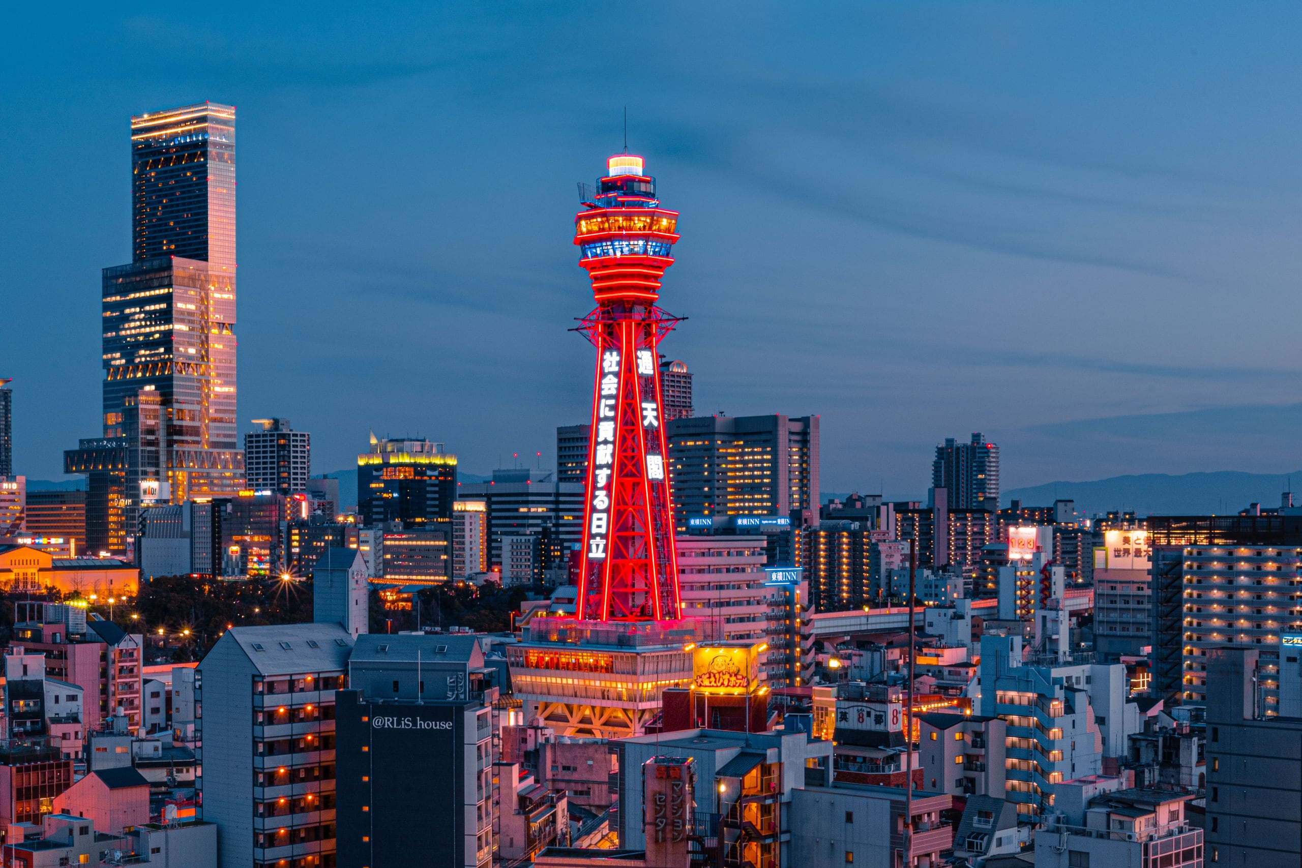 Tsutenkaku lit red at dusk above the low roofs of Shinsekai, the towers of Osaka rising behind it