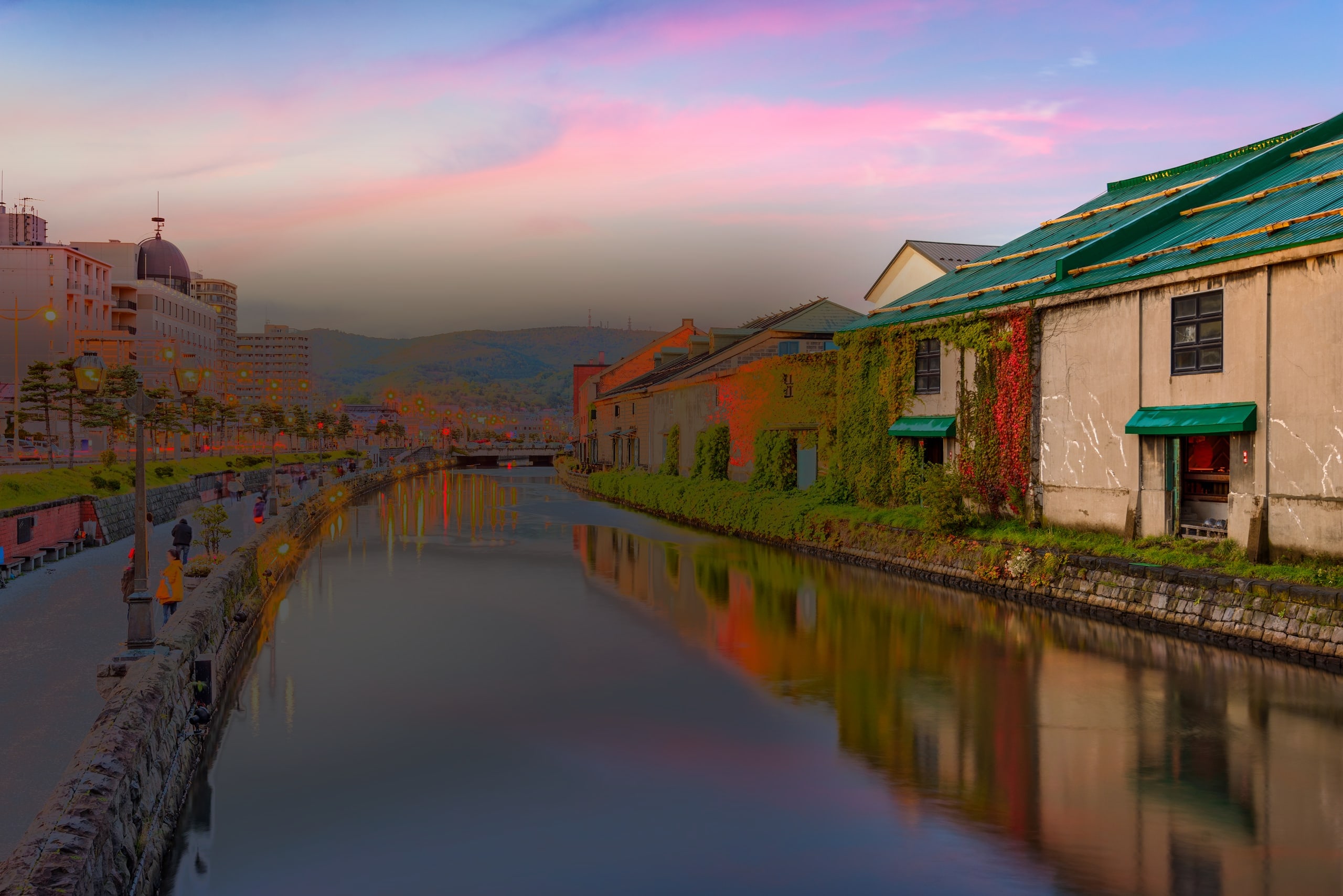 The Otaru canal at dusk under a pink sky, gas lamps lit along the water and the old stone warehouses on the far bank