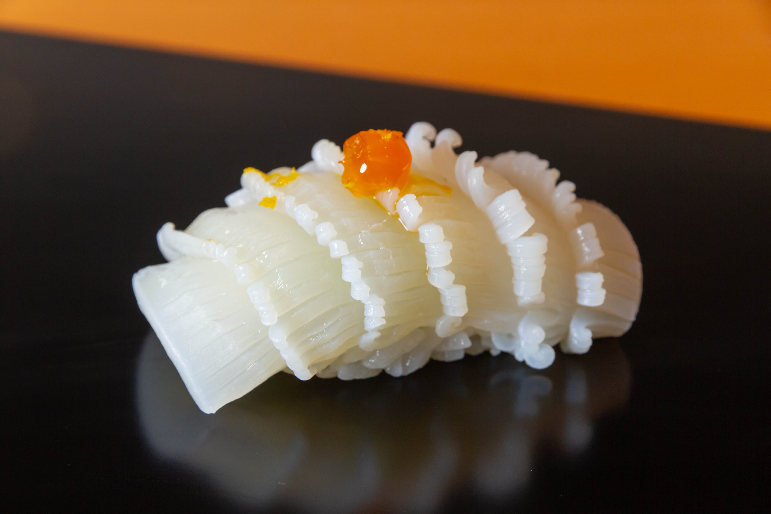 A single piece of squid nigiri, finely scored and topped with a bead of salmon roe, on a dark lacquer counter