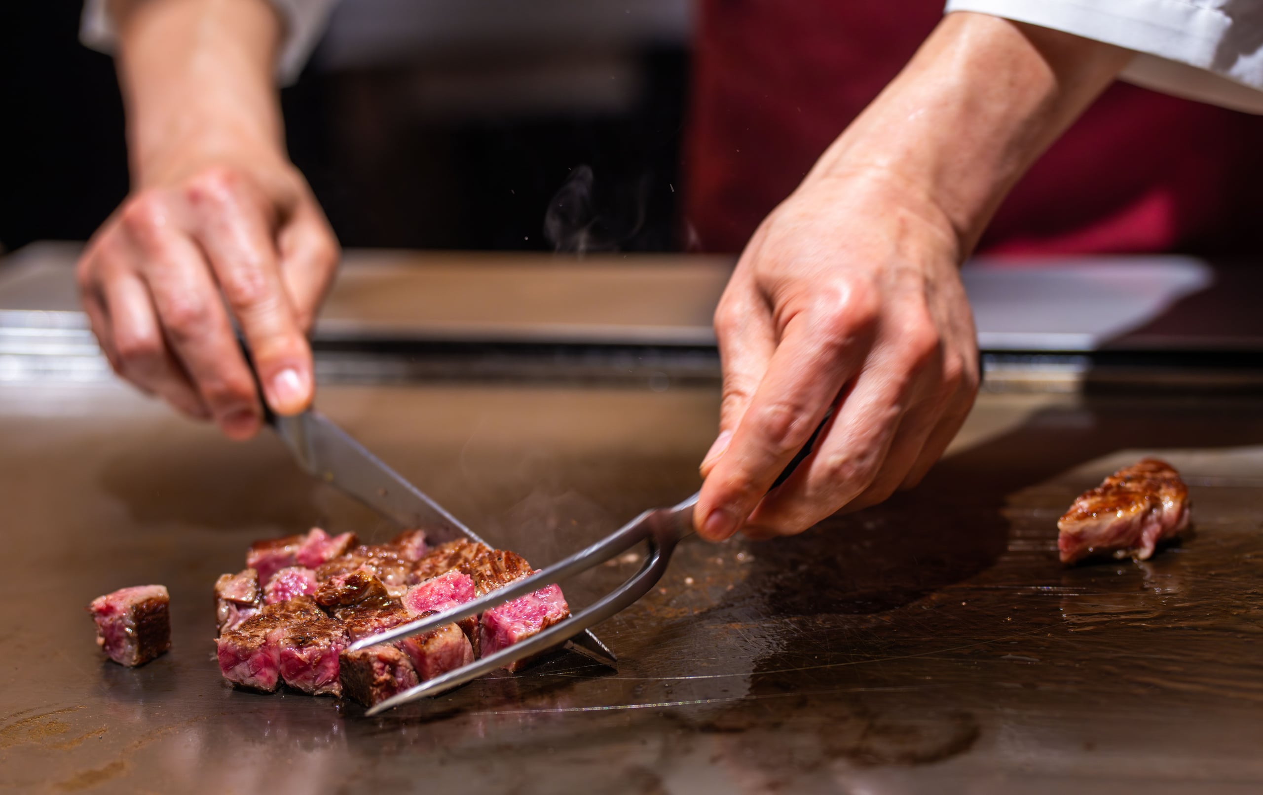 A teppan chef's hands cutting seared wagyu with tongs and a spatula, smoke rising from the plate