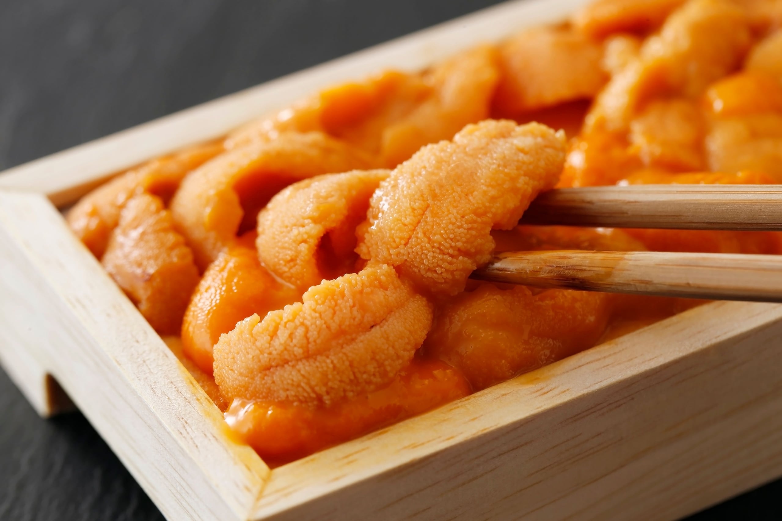 Pieces of sea urchin sashimi in a wooden box, chopsticks lifting one