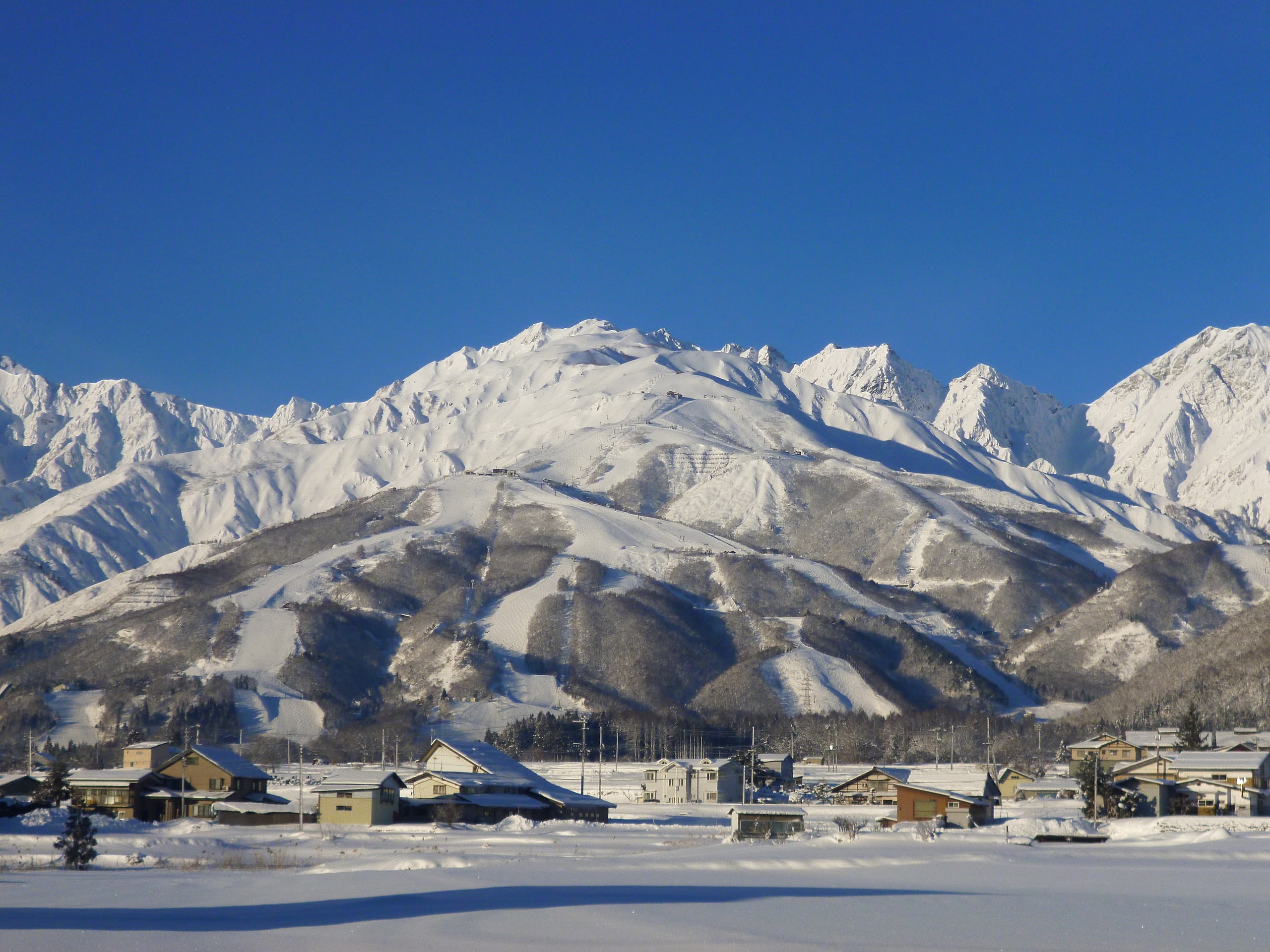 The snow-covered peaks of the Northern Alps under a deep blue sky, above the snowfields and roofs of the Hakuba valley