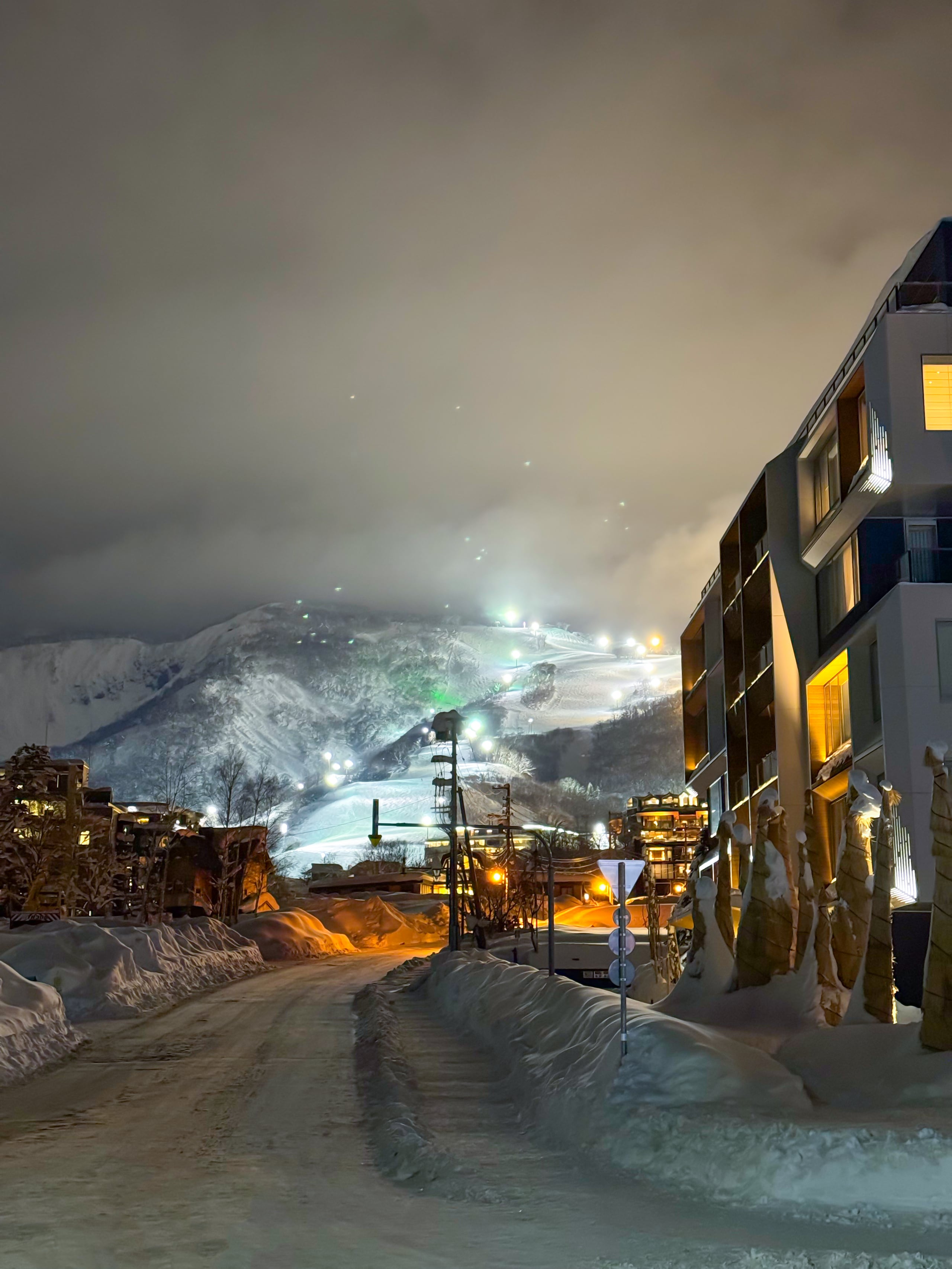 A snow-banked village street at night, lit buildings on one side and floodlit ski runs on the mountain behind