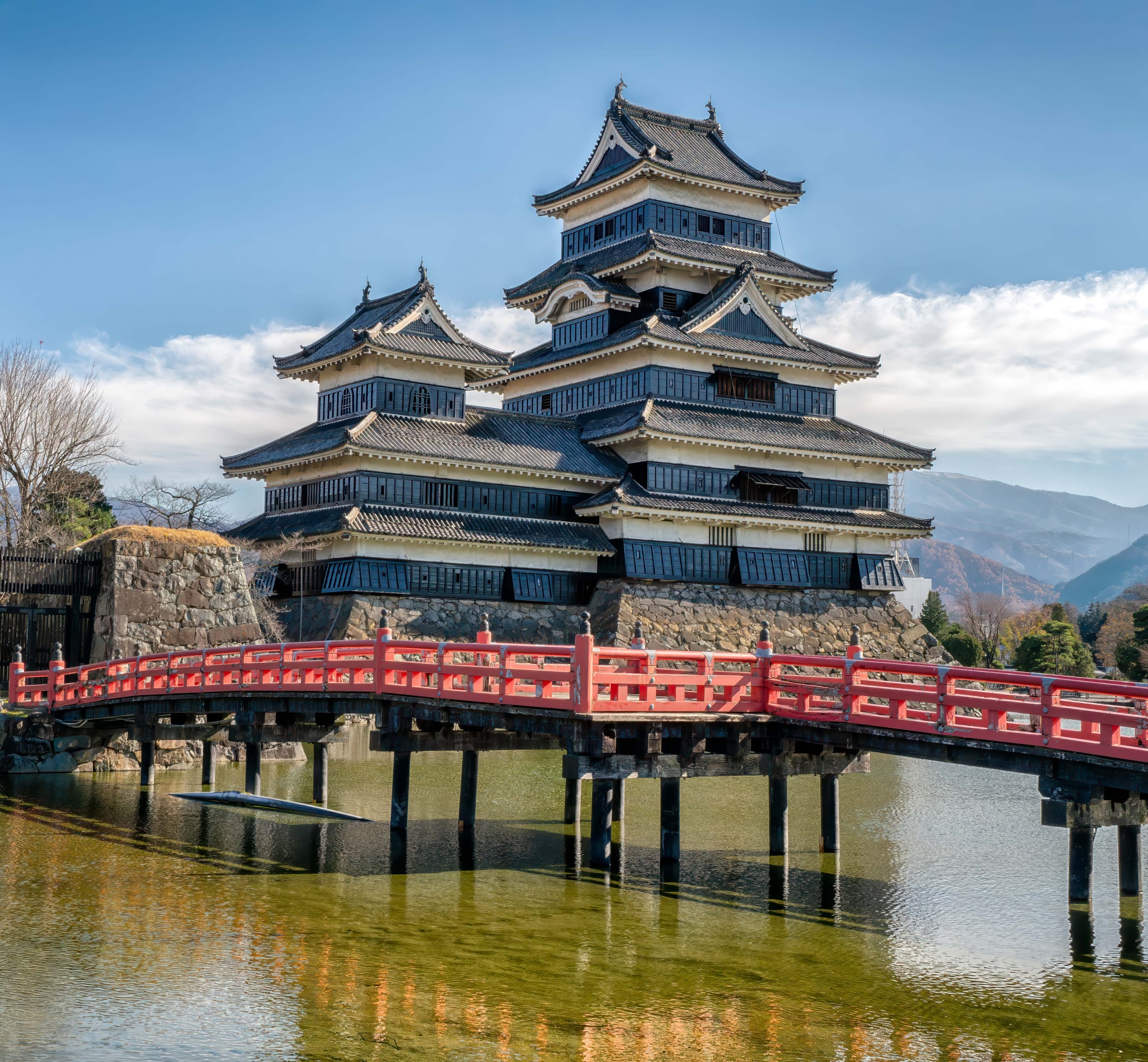 The black and white keep of Matsumoto Castle above its moat and a red bridge, snow on the roofs and the mountains behind