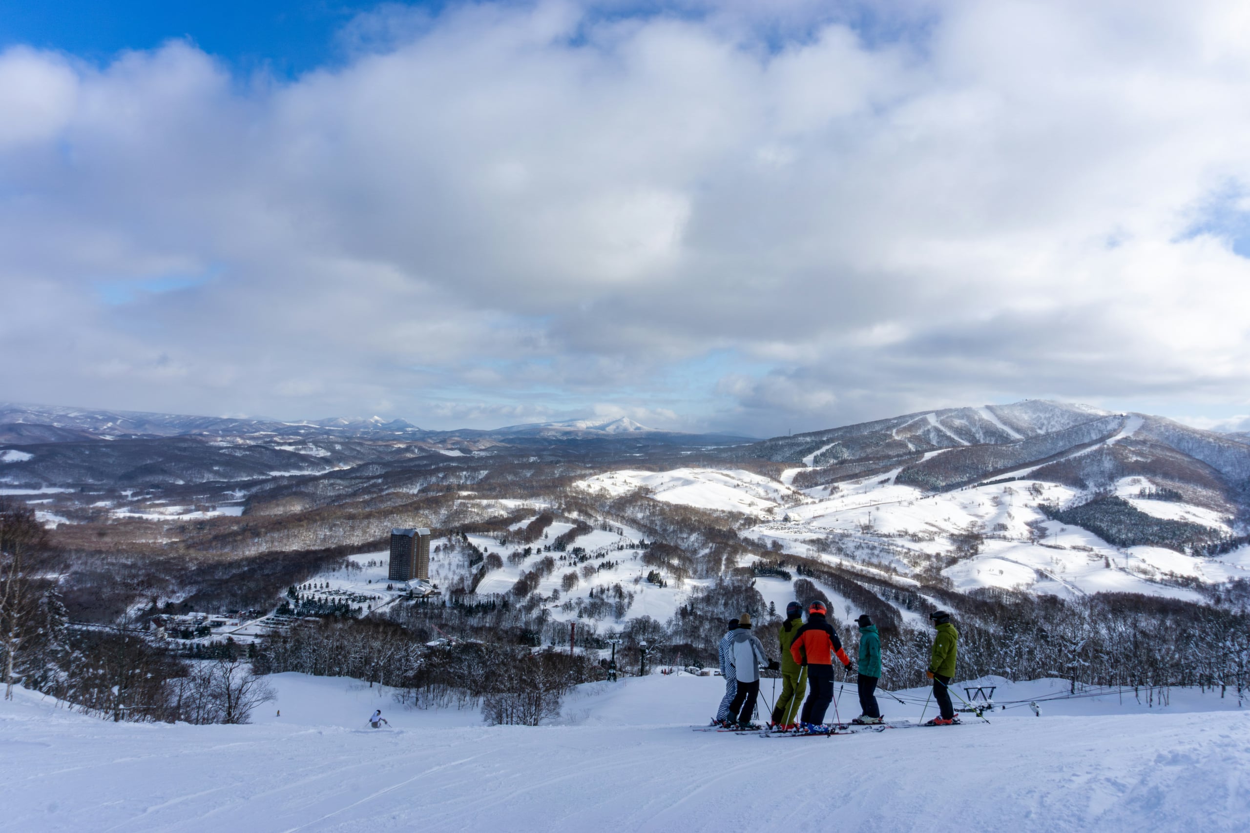 Four skiers on a ridge above Rusutsu, the resort's tower and its snow-covered mountains spread across the valley below