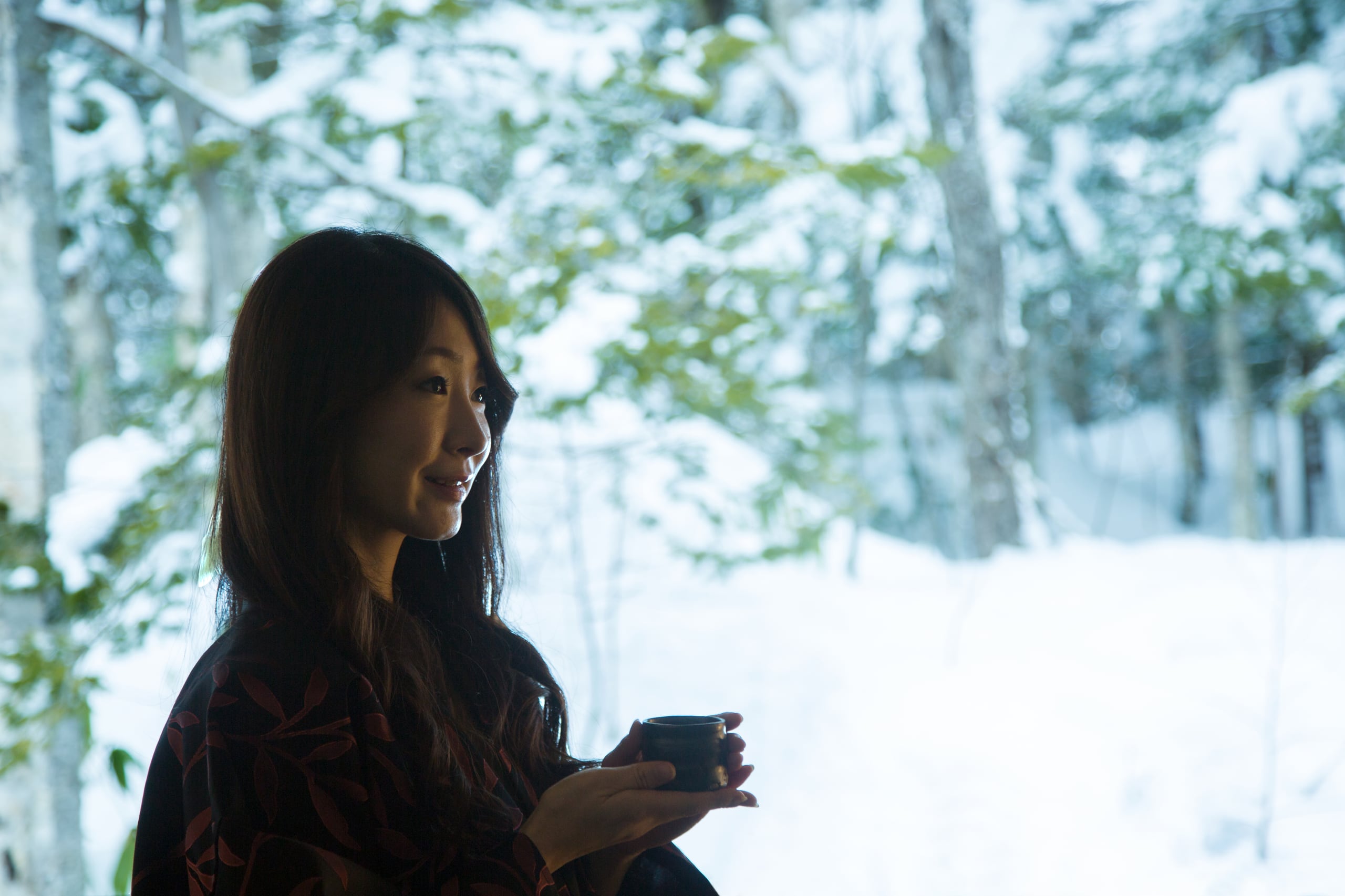 A woman in a dark yukata holding a cup of tea by a window, a forest of snow-laden birches behind her
