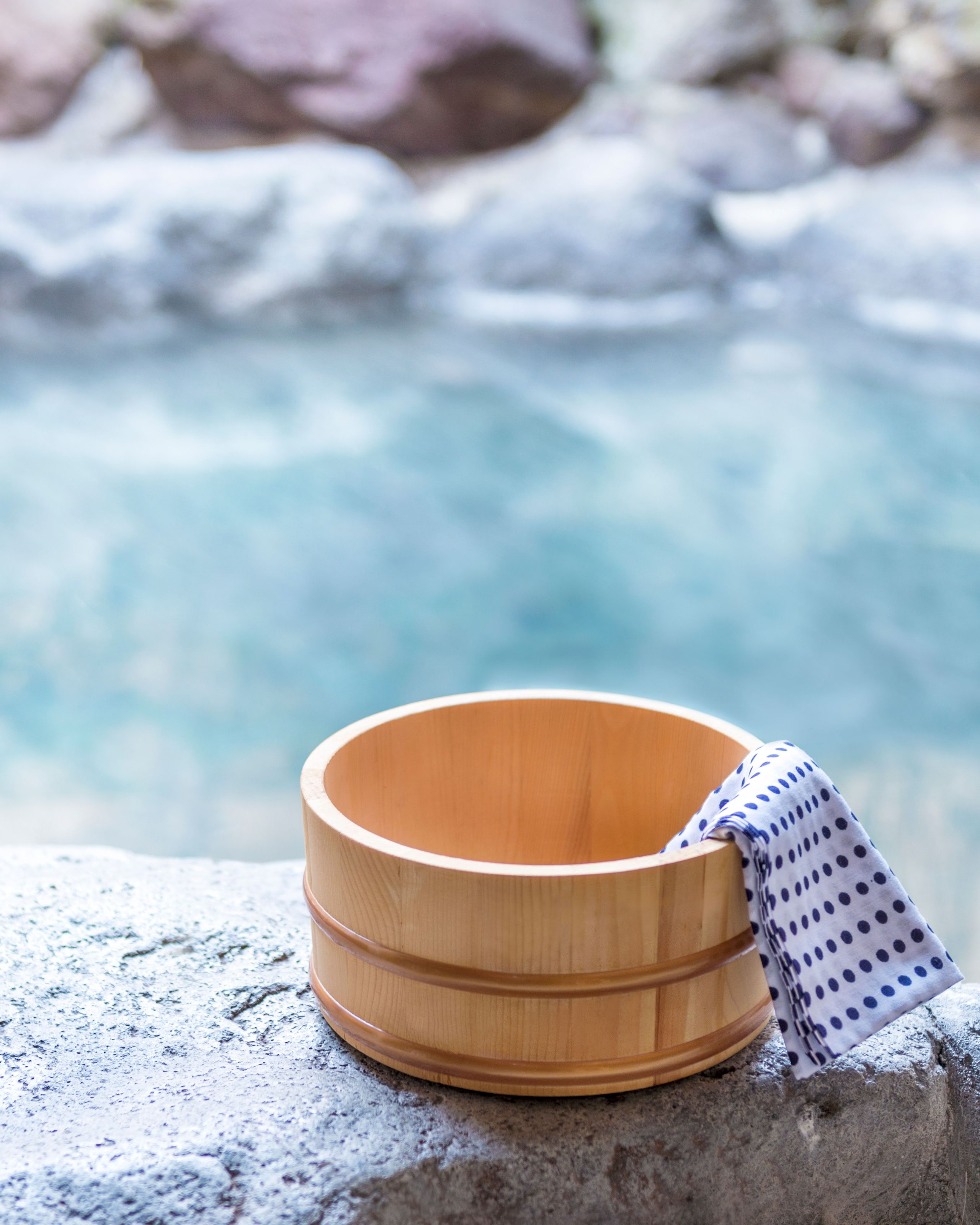 A wooden bathing pail and a blue-spotted cloth on frost-white stones beside the milky blue water of an open-air onsen