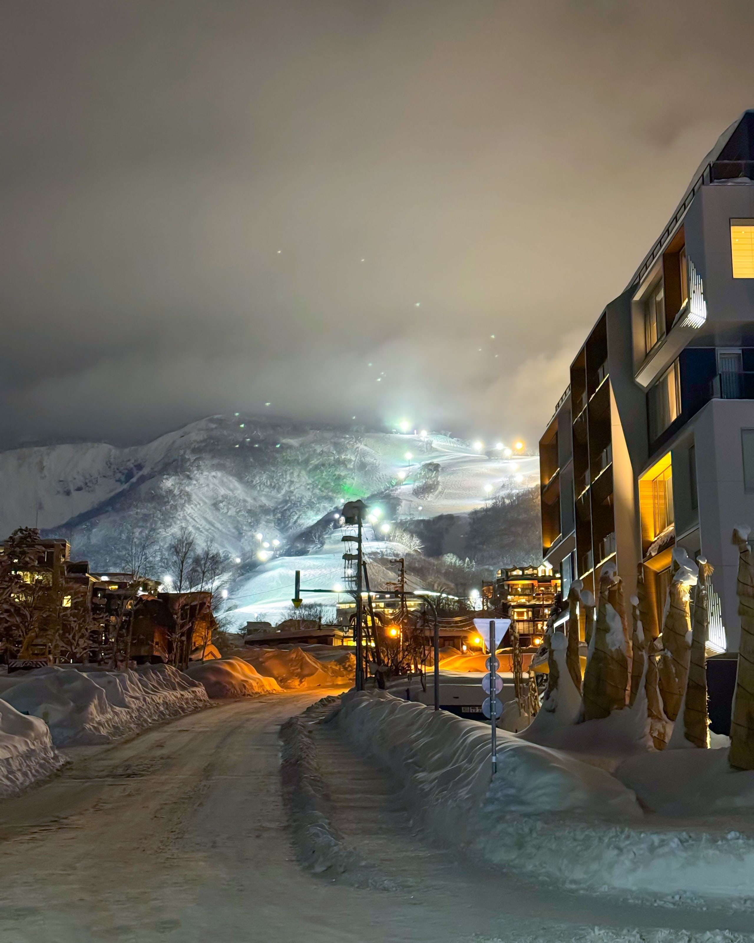 A snow-banked village street at night, lit buildings on one side and floodlit ski runs on the mountain behind