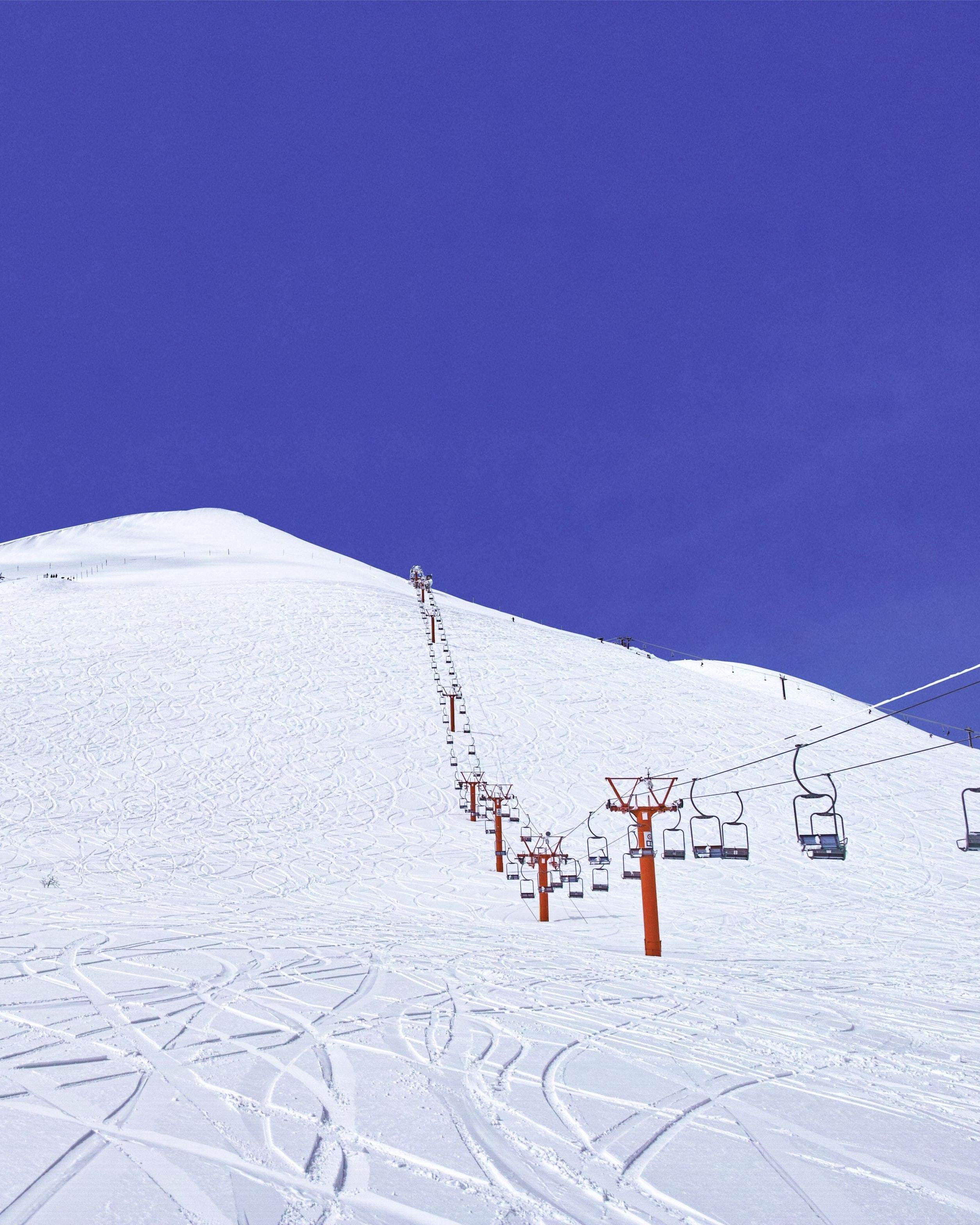 Ski tracks curving across a broad slope of fresh snow, a chairlift climbing to a rounded white summit under a deep blue sky
