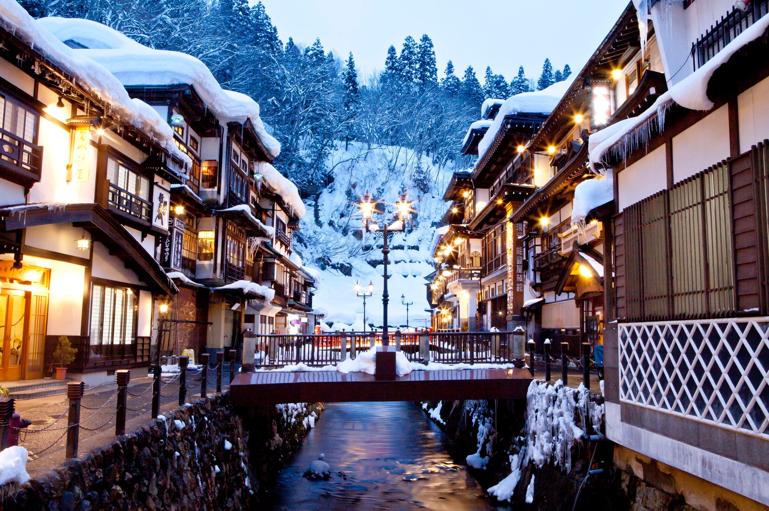 The wooden ryokan of Ginzan Onsen facing each other across a narrow river, their roofs deep in snow and their lamps lit at dusk