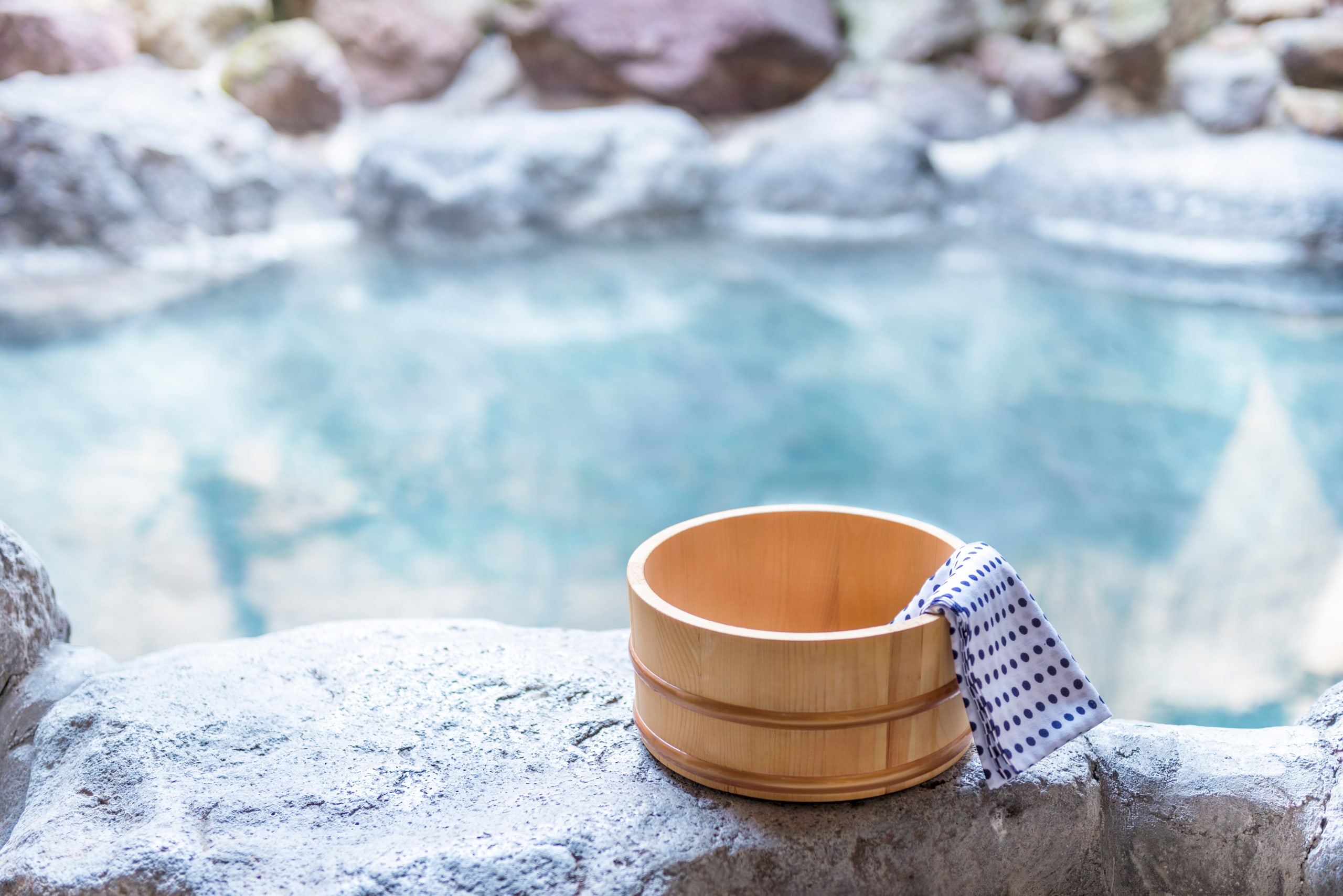 A wooden pail and a blue and white cloth on the rock edge of an open-air onsen, frost on the stones around the water