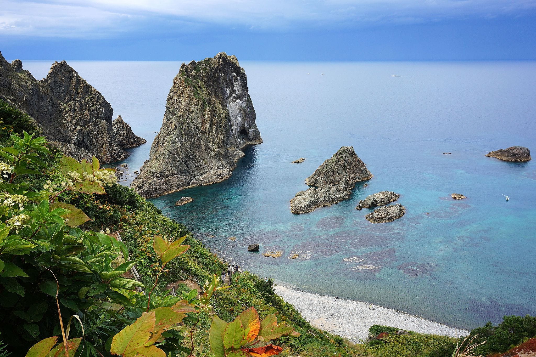 A tall sea stack and low rocks in clear turquoise water below green cliffs at Cape Kamui, Shakotan