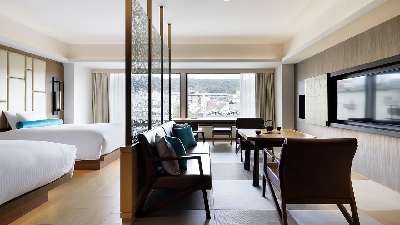 A junior suite at Hotel The Celestine Kyoto Gion, twin beds and a sofa beside a screen of woven branches, the rooftops of Kyoto beyond the window