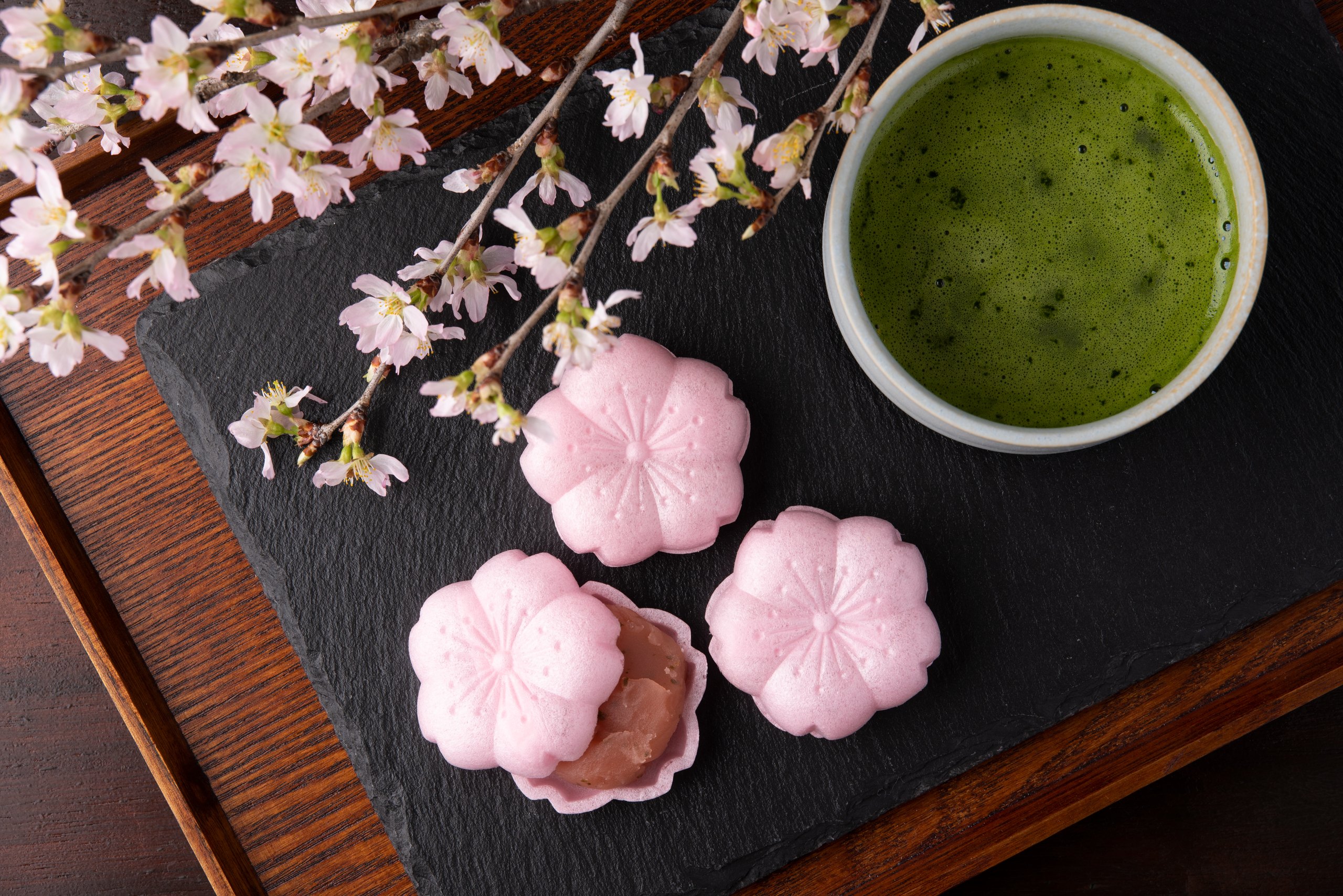 Three flower-shaped sakura monaka on a slate tray beside a bowl of whisked matcha, cherry blossom above