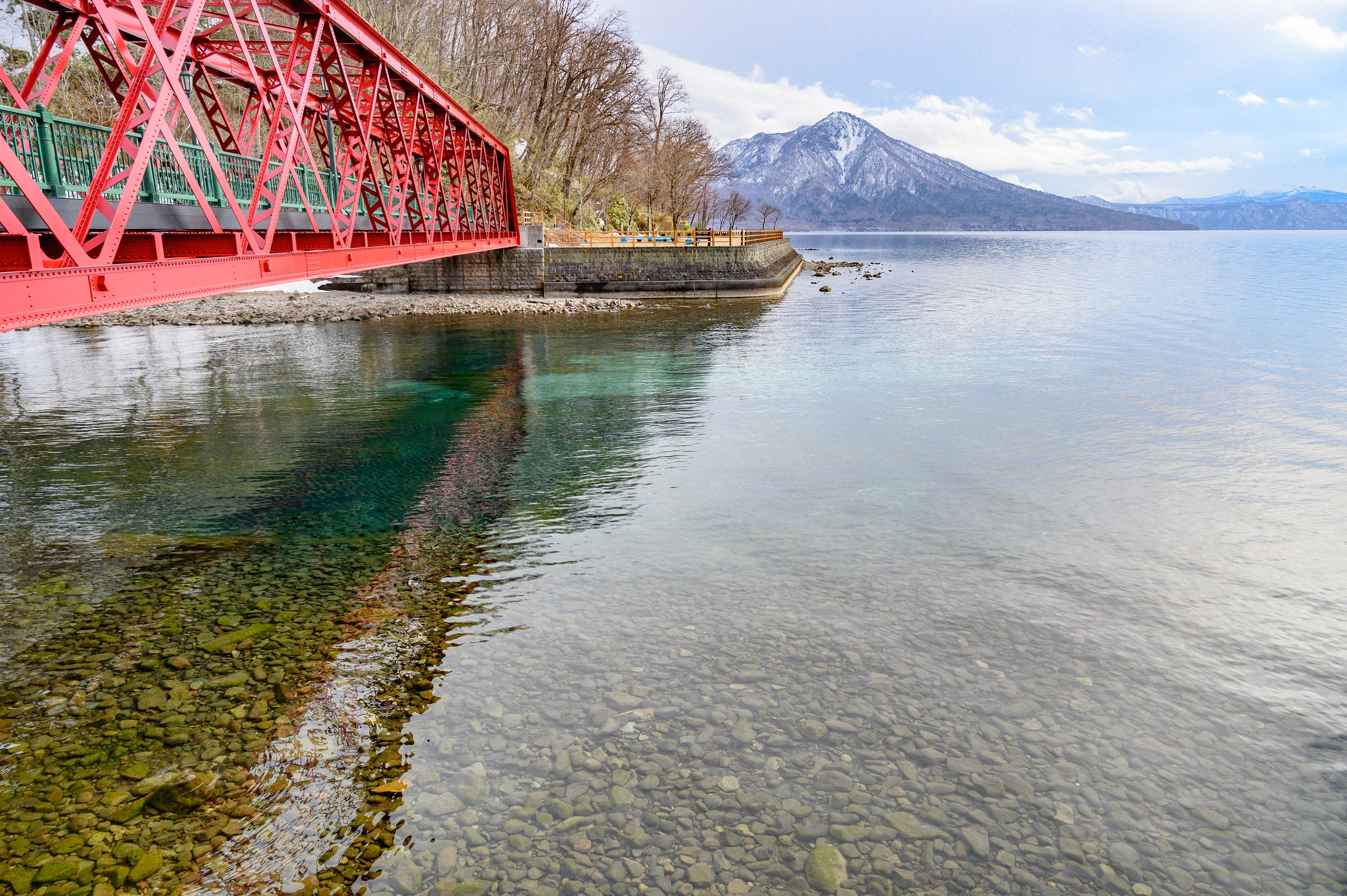 A red steel bridge at the edge of Lake Shikotsu, the stones clear beneath the water and a snow-capped peak across the lake