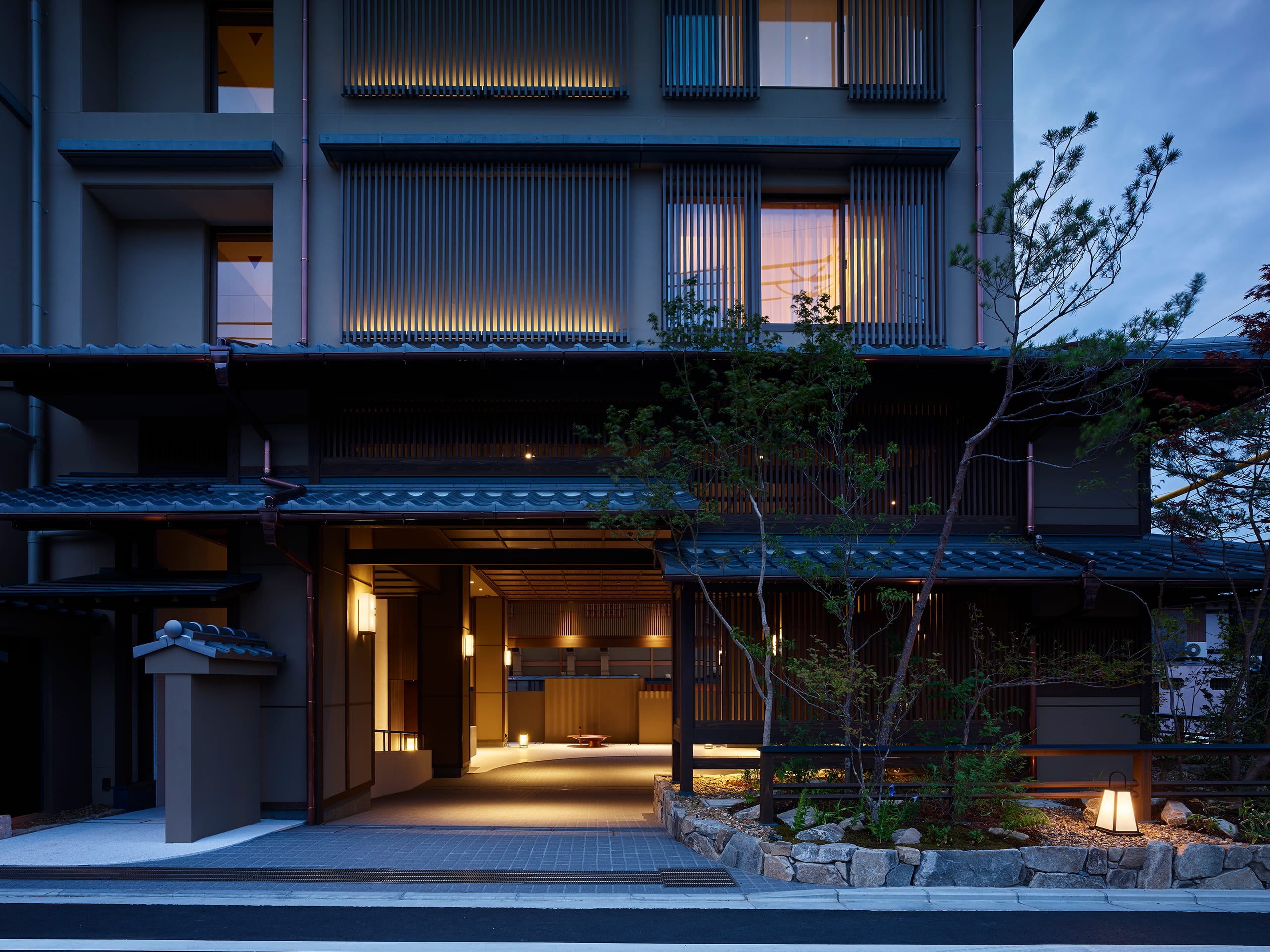 The entrance of Hotel The Celestine Kyoto Gion at dusk, timber lattice lit from within, a lantern in a stone-edged garden of slender trees