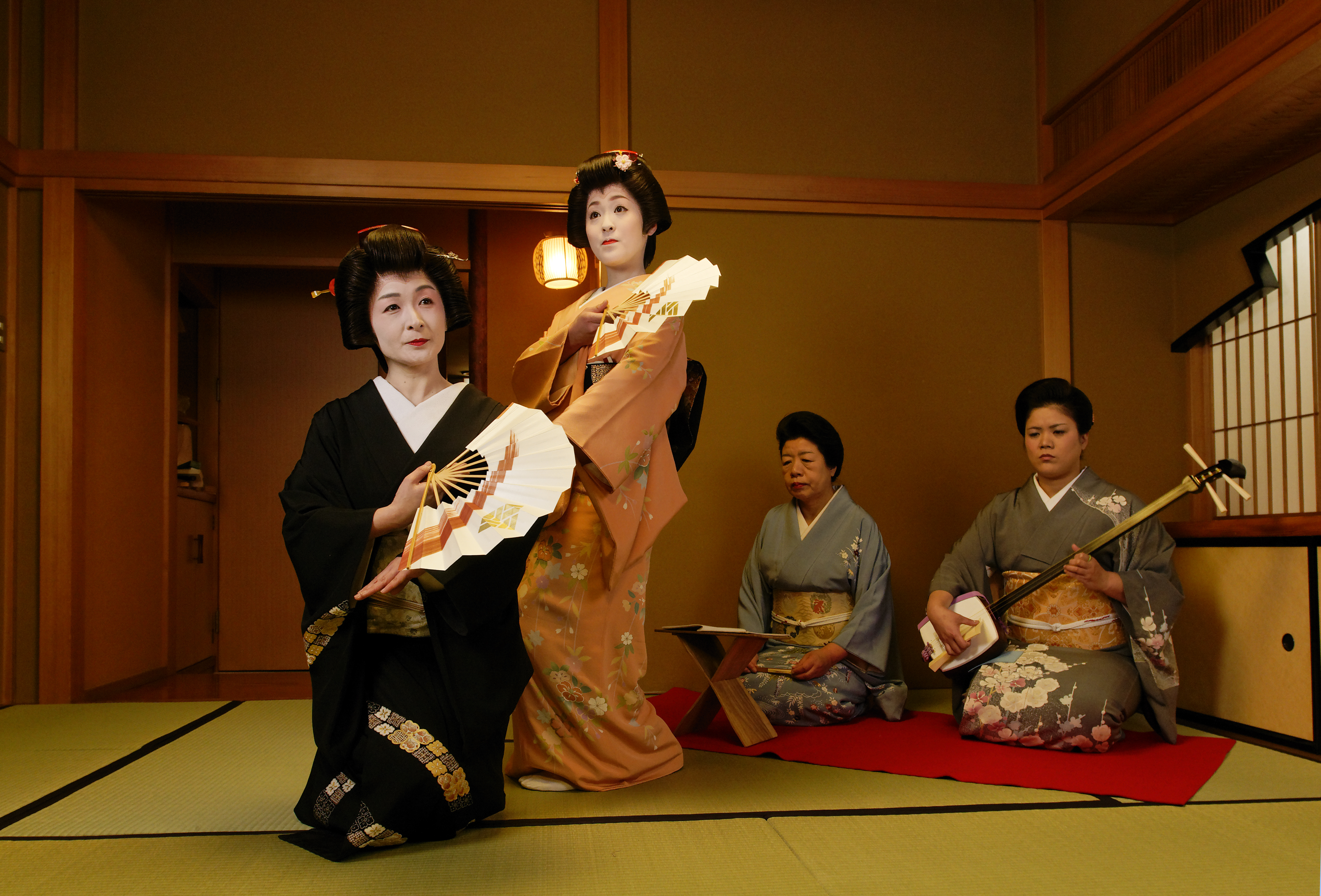 Two geiko performing a fan dance in a tatami room, accompanied by a shamisen player