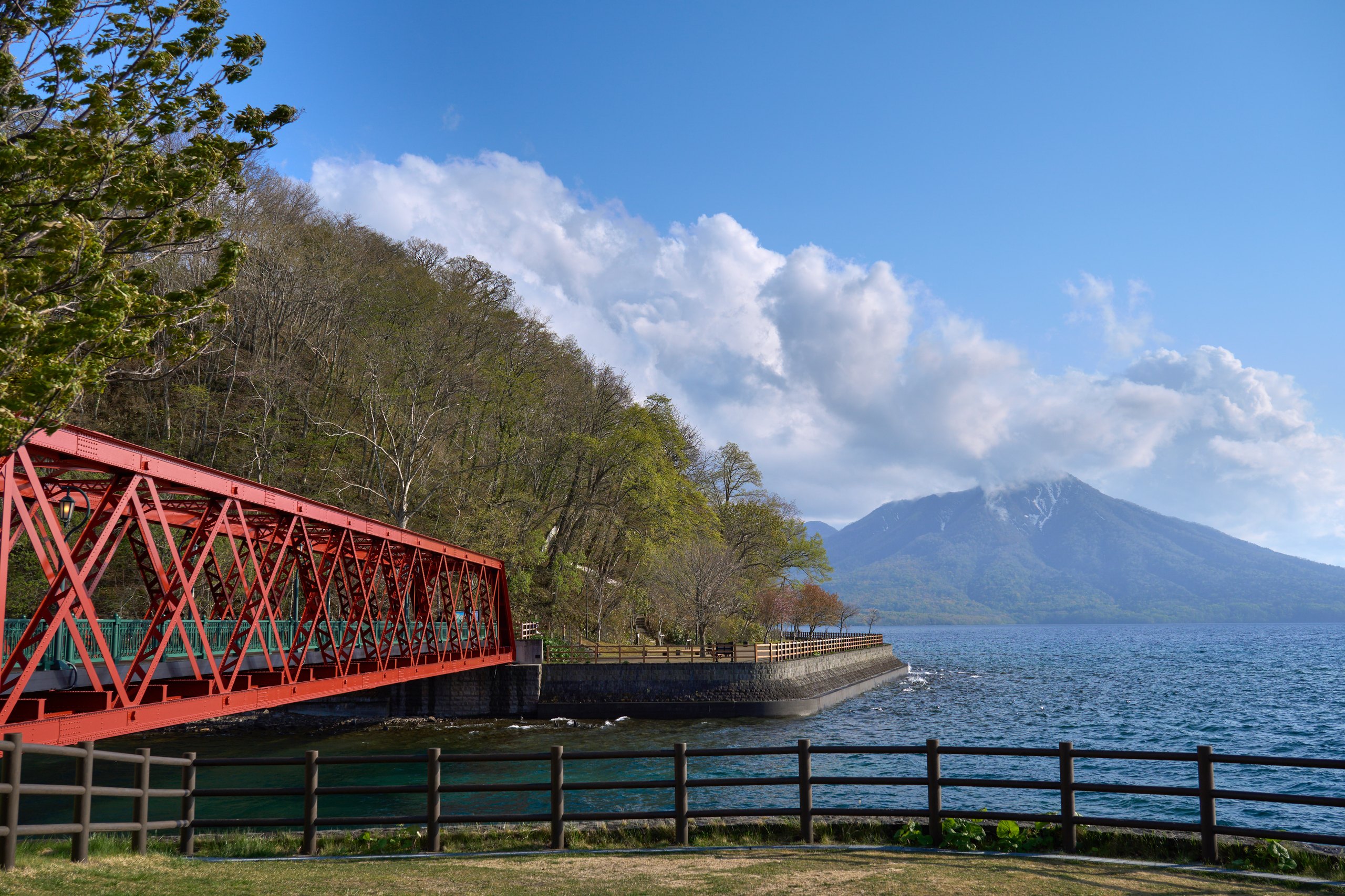 A red truss bridge on the shore of Lake Shikotsu, a volcano across the clear water