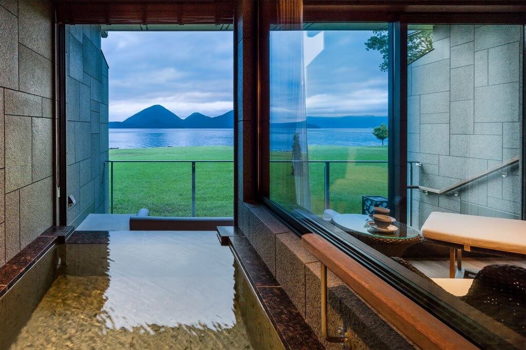 An onsen in a ground-floor suite at The Lake Suite Konosumika, the lawn and Lake Toya beyond the glass